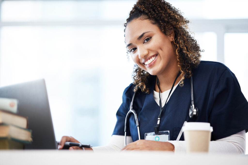 a smiling medical professional with stethoscope, laptop, pen and coffee